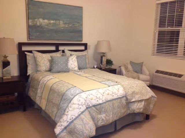 A neatly made bed with a patterned comforter and multiple pillows in a softly lit bedroom. There are two bedside tables with lamps on each side of the bed. A framed painting hangs on the wall above the headboard. To the right, there is a white armchair with a cushion and a window with blinds. Below the window is a heating or air conditioning unit.