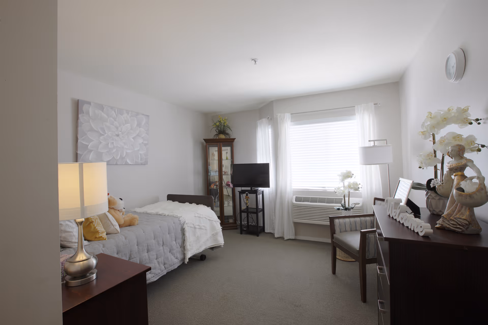 A cozy, well-lit bedroom in a senior living facility featuring a single bed with a gray quilt and a teddy bear, a bedside table with a lamp, a glass display cabinet, a flat-screen TV on a stand, a window with white curtains and an air conditioning unit, a wooden chair, and a dresser decorated with flowers, a 'Welcome' sign, and a figurine.