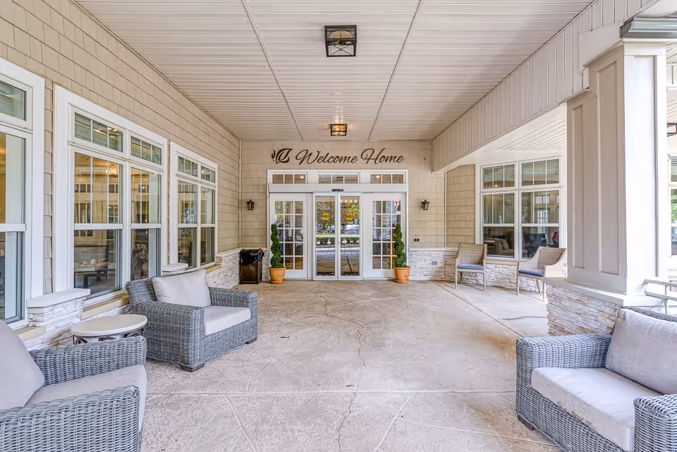 Covered entrance area of Chesapeake Place Senior Living with wicker chairs and small tables arranged on both sides. The walls have multiple windows, and a double glass door is centered under a sign that reads 'Welcome Home'. Two potted plants flank the entrance doors.