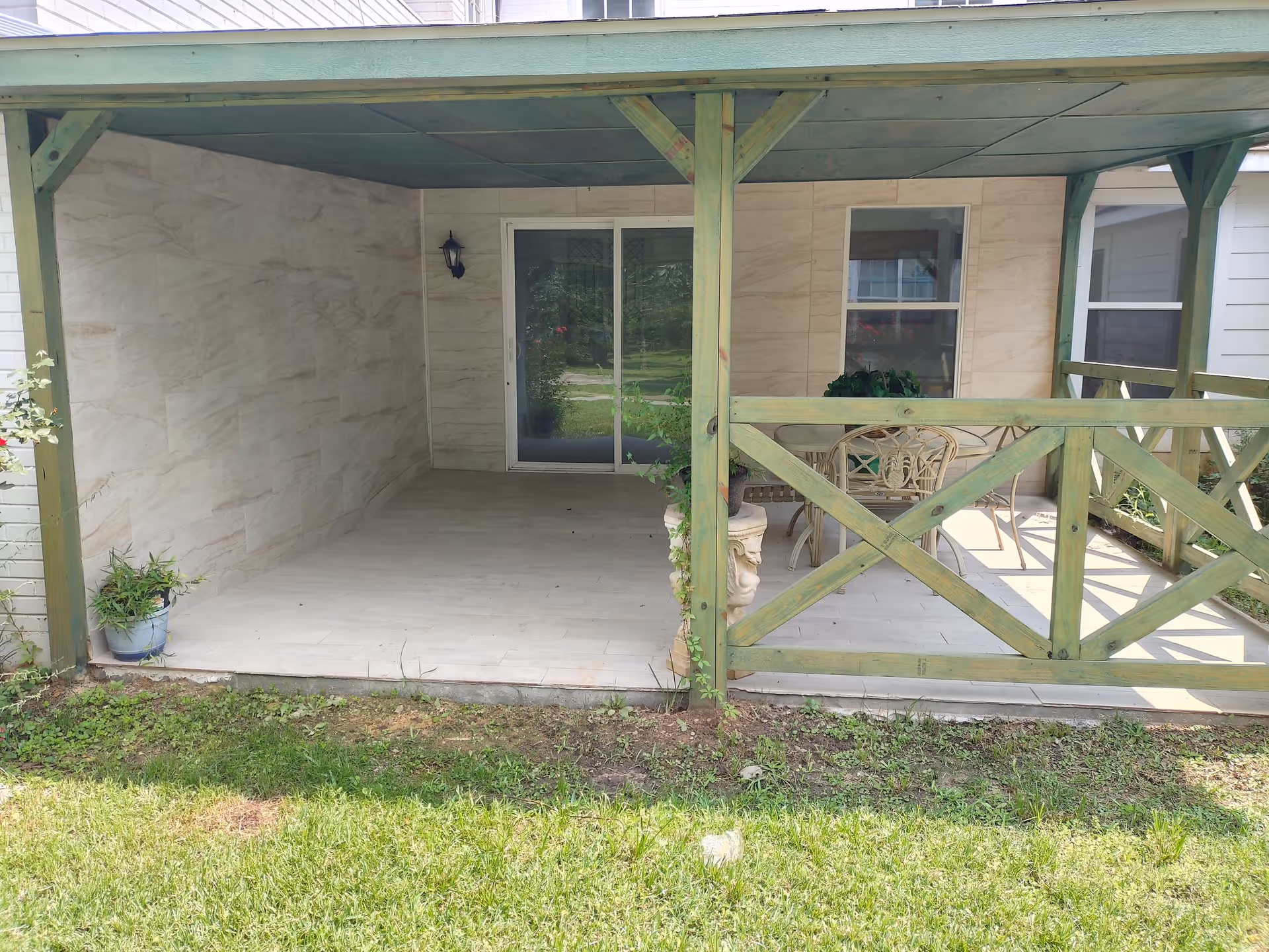 Covered outdoor patio area with a green wooden railing and support beams. The patio has tiled flooring and a small seating area with a table and chairs. There are potted plants placed on the patio and grass in the foreground.