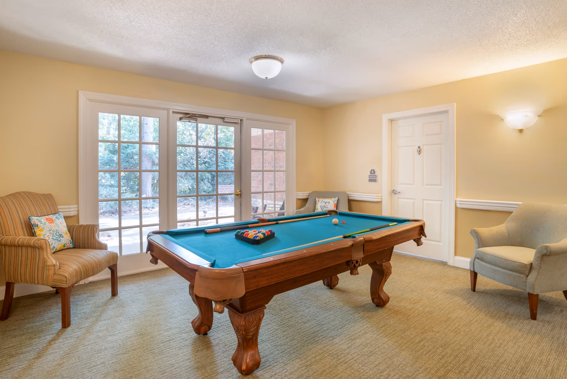 A cozy indoor room with a pool table in the center, surrounded by three upholstered armchairs. Behind the pool table, there are large glass doors showing an outdoor view with trees. The walls are painted light yellow and the room is softly lit by ceiling and wall lights.
