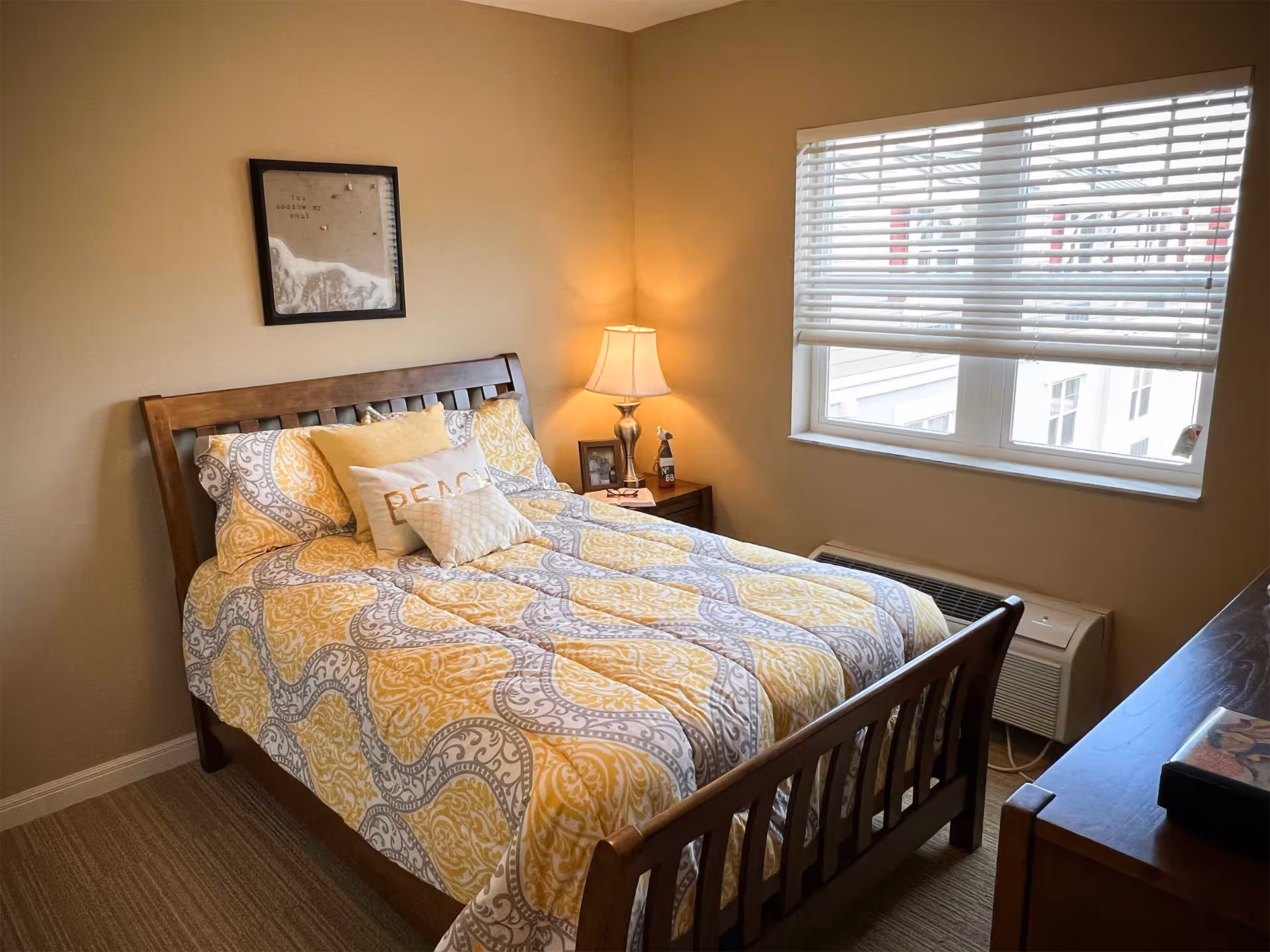 A cozy bedroom with a wooden bed frame and a bed made with a yellow and gray patterned comforter and pillows, including one with the word 'PEACE'. A nightstand with a lamp and picture frames is beside the bed. A window with white blinds lets in natural light, and a framed picture hangs on the beige wall above the bed.
