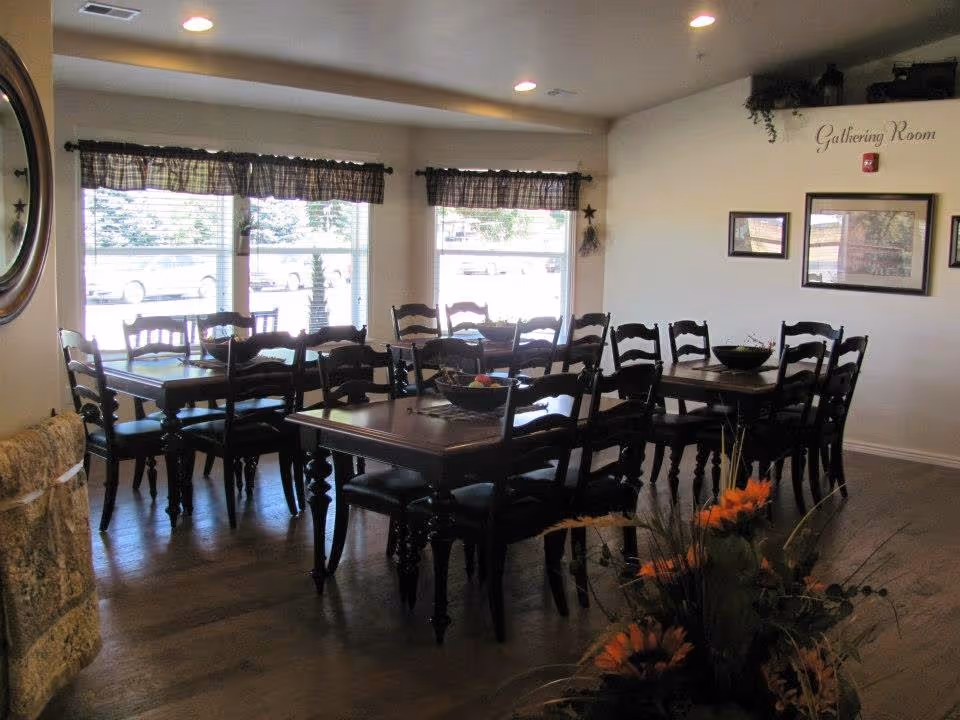 Interior view of a gathering room with multiple dark wooden tables and chairs arranged neatly. Large windows with checkered valances allow natural light to fill the room. The walls are decorated with framed pictures and the words 'Gathering Room' are written on one wall. A floral arrangement is visible in the foreground.
