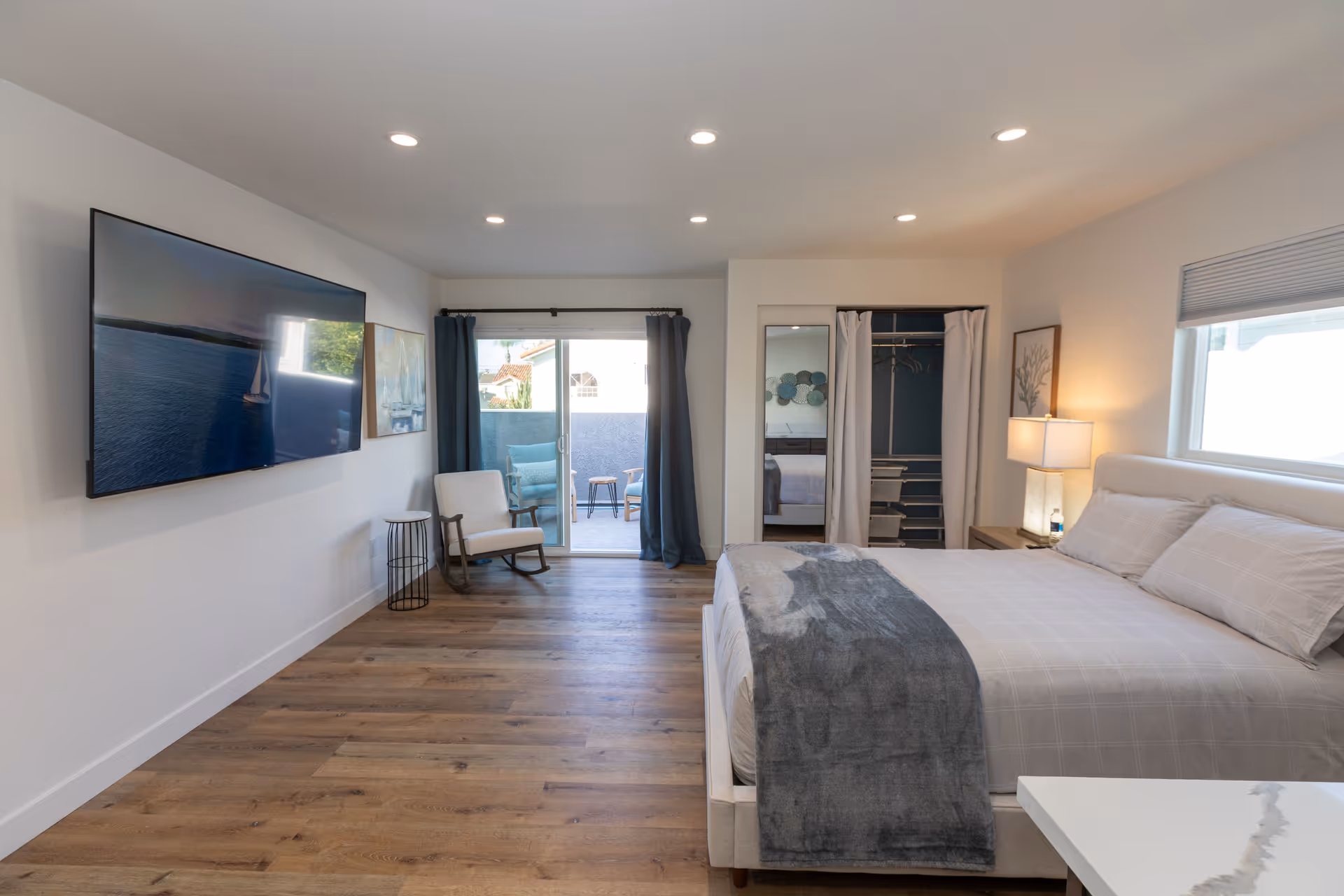 A modern bedroom with a large bed covered in light-colored bedding and a gray throw blanket. There is a nightstand with a lamp and a water bottle next to the bed. A closet with white curtains and a full-length mirror is visible. A sliding glass door with dark curtains leads to a small outdoor seating area with chairs and a table. A large flat-screen TV is mounted on the wall opposite the bed, and a rocking chair with a small side table is placed near the door. The room has wooden flooring and recessed ceiling lights.
