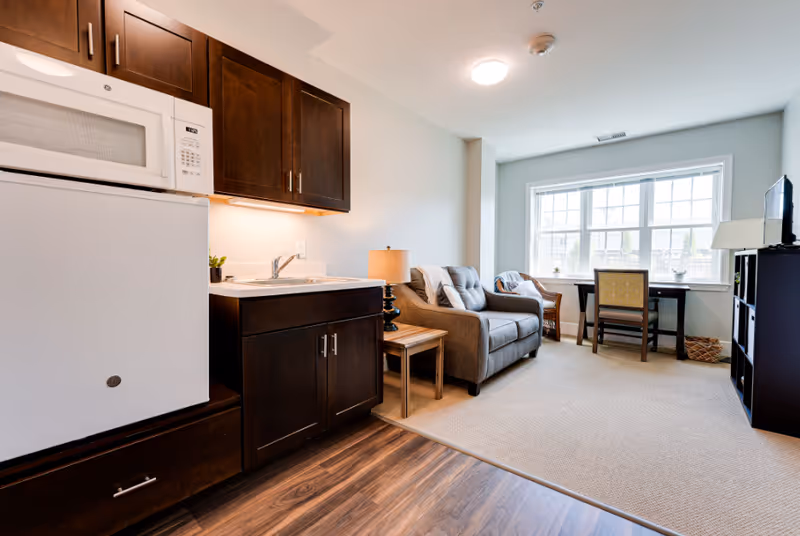 A bright and cozy living space featuring a small kitchenette with dark wood cabinets, a white microwave, and a mini refrigerator. Adjacent to the kitchenette is a sitting area with a gray upholstered loveseat, a wooden side table with a lamp, a wicker chair, and a desk with a chair positioned in front of a large window letting in natural light. A black shelving unit with a lamp and a TV is also visible.