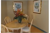 Small dining area with a round wooden table, four chairs, a floral centerpiece, and framed artwork on beige walls.