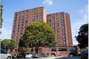 Front exterior of a tall brick senior living center with trees and parked cars.