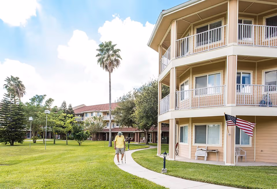 A senior man with a cane walking on a curved sidewalk through a grassy courtyard with trees and a multi-story beige building with balconies and an American flag on the right side.