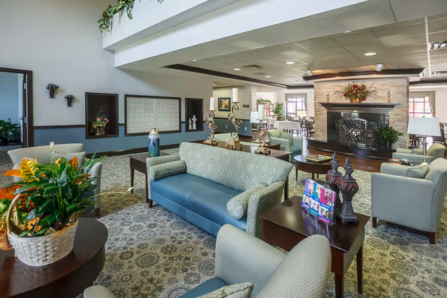 A spacious and well-lit living room area in a senior living facility featuring comfortable upholstered sofas and armchairs arranged around a stone fireplace. The room has patterned carpeting, decorative plants, and various small tables with decorative items. Mailboxes and restroom signs are visible on the wall in the background.