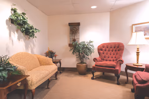 A cozy sitting area with a beige patterned sofa, a red upholstered armchair, and a matching red ottoman. There are wooden side tables with a lamp and decorative plants, including a wall-mounted plant and a potted plant on the floor. The walls are light-colored, and there is a framed picture and a decorative wall piece.