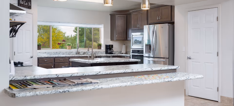 Modern kitchen with granite countertops, a central island with a sink, stainless steel refrigerator, built-in microwave and oven, coffee maker, and large window overlooking greenery. Magazines are placed on the extended countertop bar area.