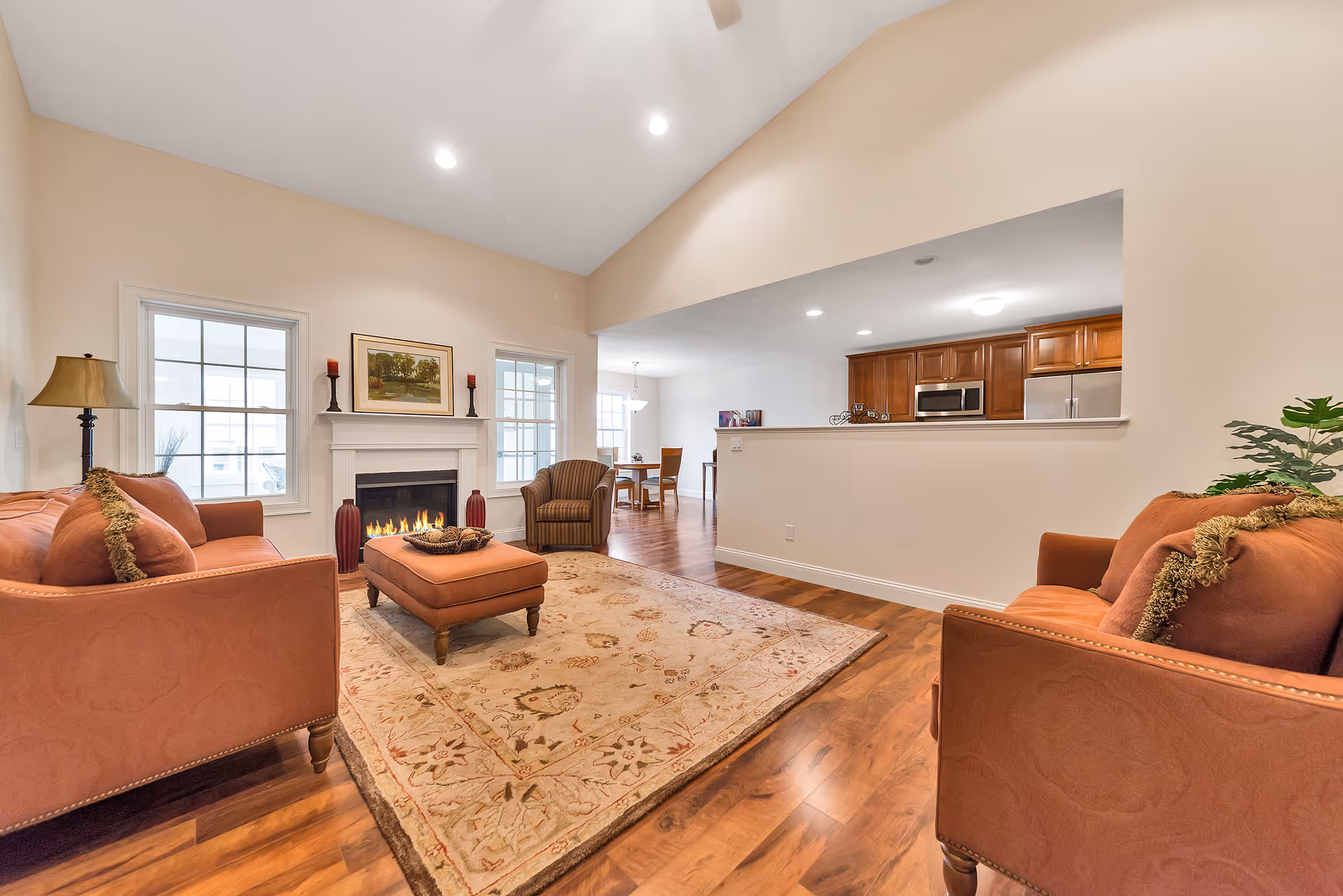 A cozy living room with two orange sofas, a matching ottoman, a patterned area rug, a fireplace with a painting and candles on the mantel, and a view into a dining area and kitchen with wooden cabinets and stainless steel appliances.