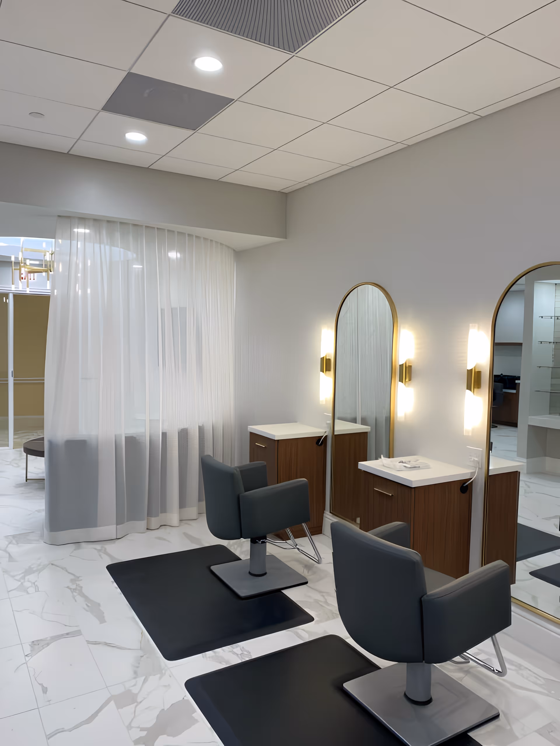 Interior view of a modern salon area with two black salon chairs placed on black mats in front of two large arched mirrors mounted on a light gray wall. Each mirror has two vertical wall lights on either side. There are wooden cabinets with white countertops beneath the mirrors. The floor is covered with white marble tiles, and a sheer white curtain partially separates this area from another space in the background.