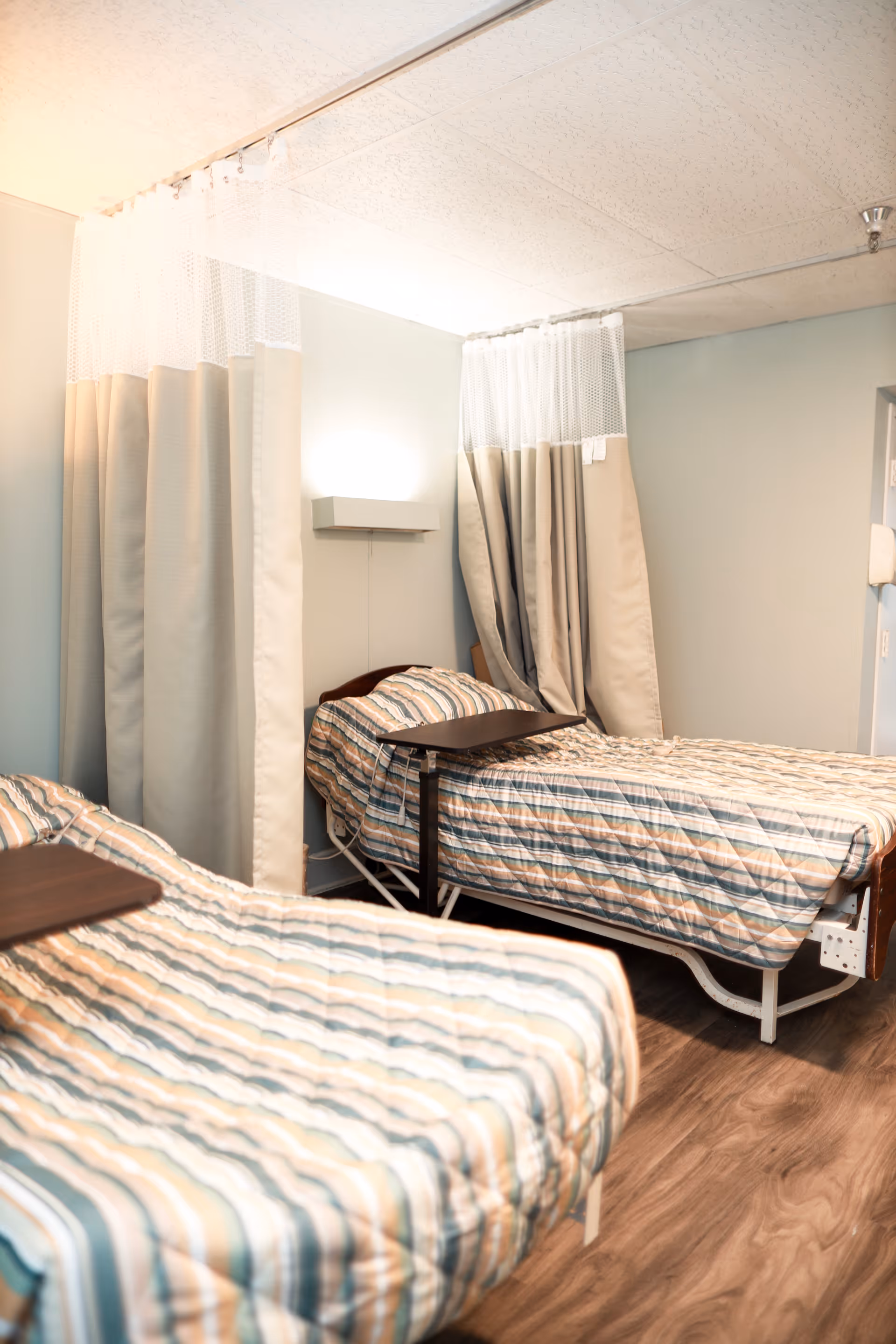 A room with two hospital-style beds separated by privacy curtains. Each bed has a striped bedspread and an overbed table. The room has light-colored walls and wood flooring.