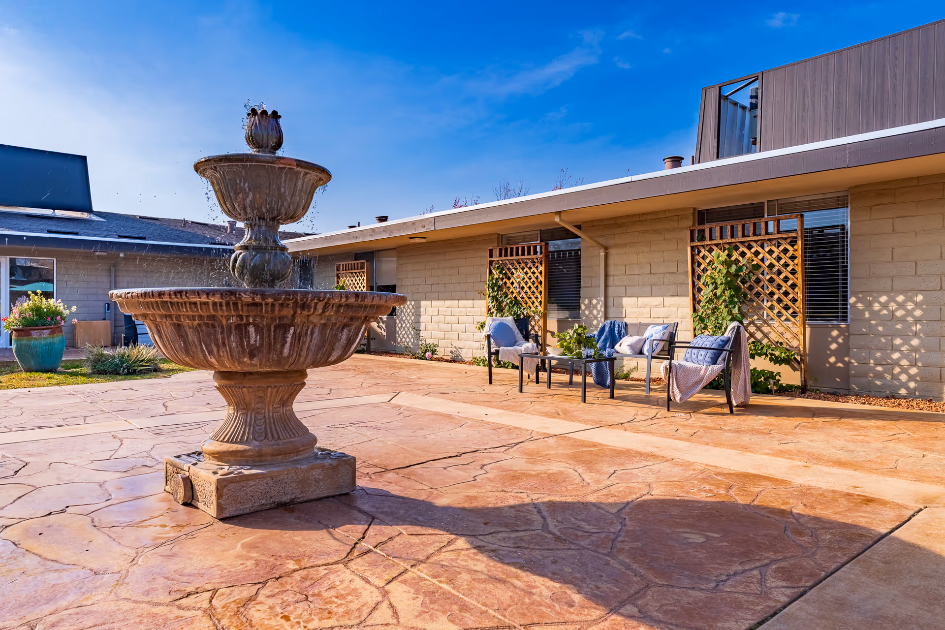 Sunlit stone courtyard featuring a tiered fountain in the foreground and a seating area with chairs and trellises against a single-story building.