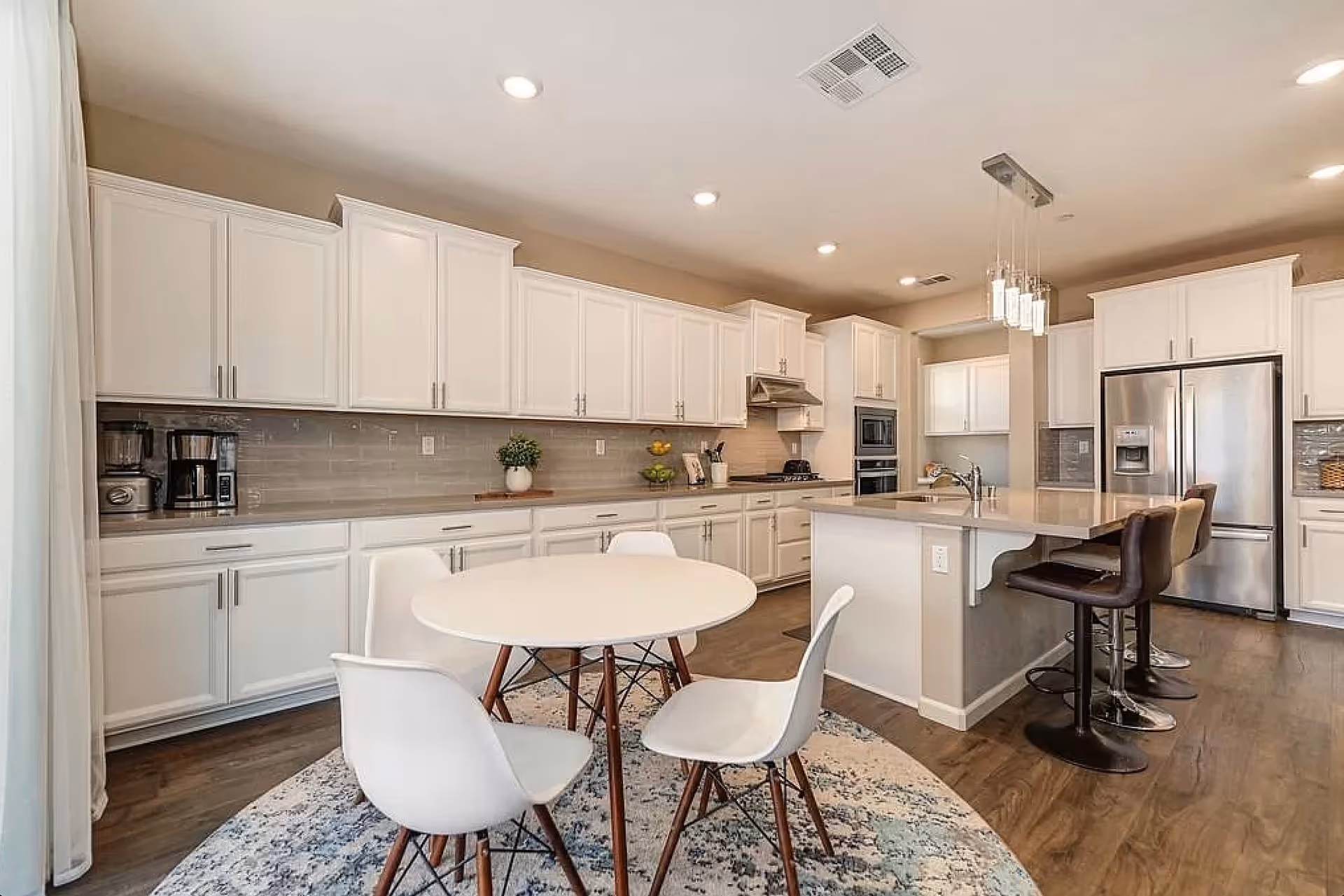 A modern kitchen with white cabinetry, stainless steel appliances including a refrigerator and built-in oven, a kitchen island with three brown bar stools, and a round white dining table with four white chairs on a patterned rug. The room has recessed lighting and a hanging light fixture above the island.