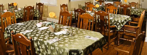 Dining room with multiple wooden tables covered with green tablecloths featuring a leaf pattern. Each table is set with cups, saucers, utensils, and small flower vases with yellow flowers. Wooden chairs surround the tables and windows with blinds are visible in the background.
