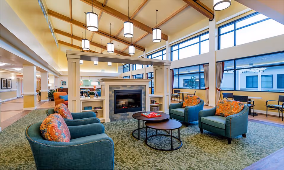 A spacious and well-lit living room area with high ceilings and large windows. The room features four teal armchairs with colorful patterned cushions arranged around two round nesting coffee tables. A modern fireplace with built-in shelves is centrally located, dividing the space. Pendant lights hang from the wooden beam ceiling, and additional seating is visible near the windows.