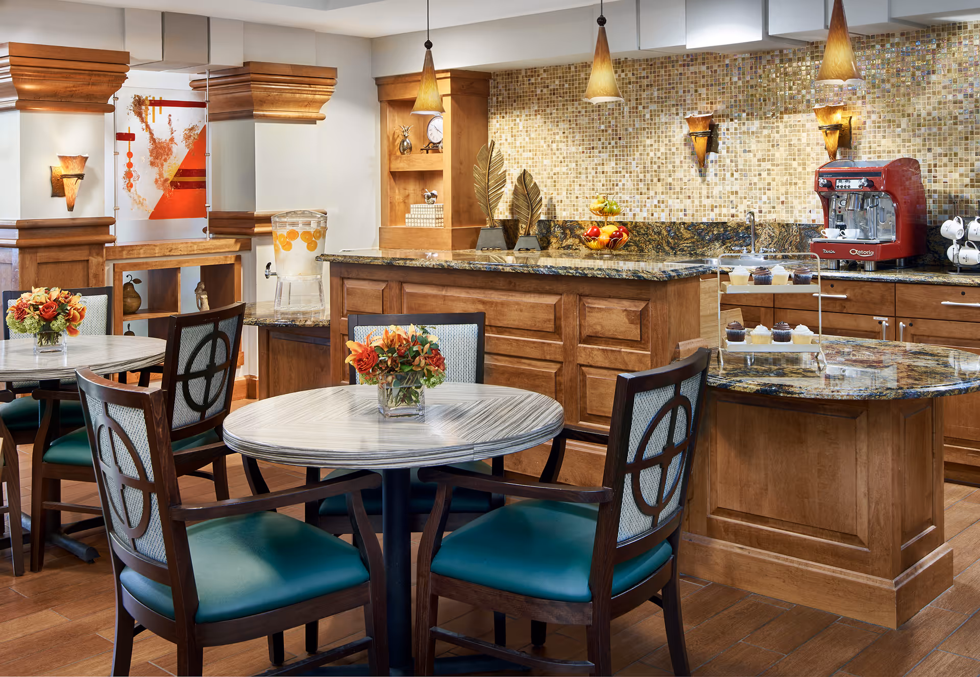 A cozy dining area with round tables and wooden chairs featuring teal cushions. Each table has a small floral centerpiece. The background shows a kitchen counter with a granite countertop, a red espresso machine, a glass dispenser with lemon water, and a tiered stand with cupcakes. The walls have mosaic tile backsplash and wooden cabinetry with decorative elements.
