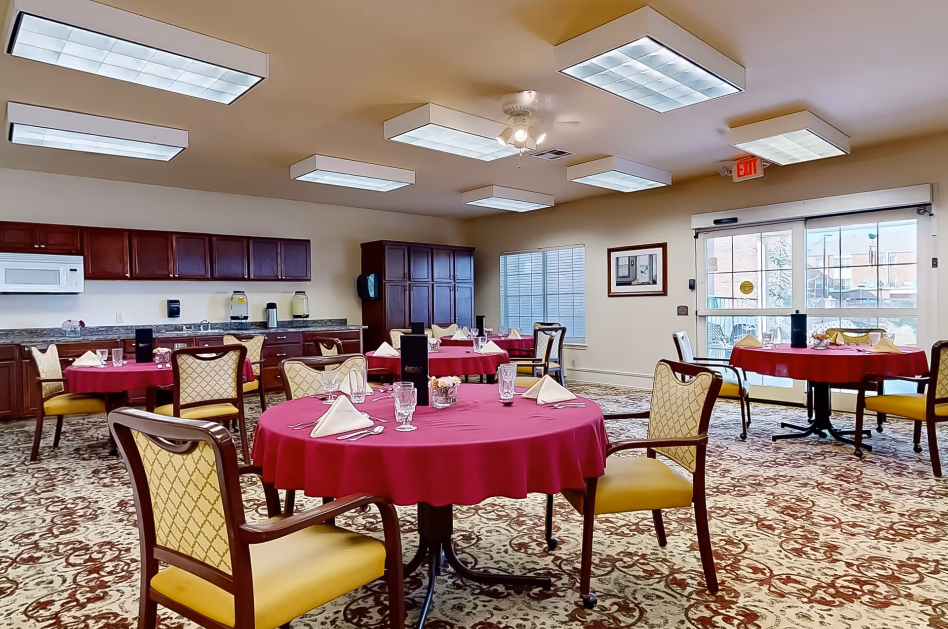 A dining room with round tables covered in red tablecloths, each set with folded napkins, glasses, and silverware. The room has patterned carpet flooring, wooden chairs with yellow cushions, and a kitchen area with dark wooden cabinets, a microwave, and a countertop in the background. Large ceiling lights and a ceiling fan illuminate the space, and there is a glass door leading outside.