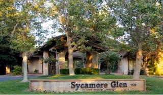 Exterior view of the Sycamore Glen Active Senior Community building surrounded by trees and greenery, with a stone sign displaying the community name in front.