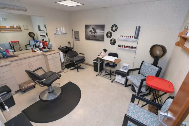A small salon room with two black salon chairs, a large mirror above a counter with various hair care products, a hair dryer chair, and a manicure station with nail polish displayed on the wall. There are also two patterned armchairs and a red tray table in the corner.
