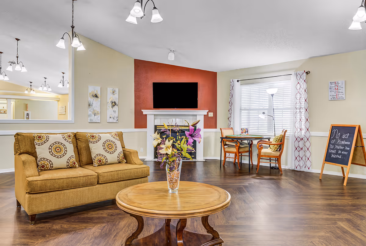 Bright communal living room with a sofa, coffee table, fireplace and TV, and a small table and chairs by a window.