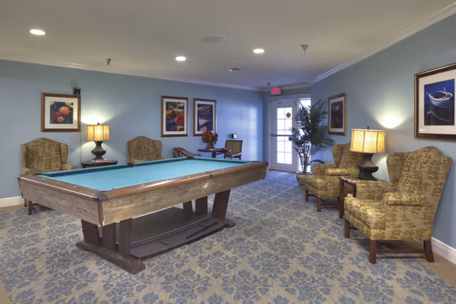 A well-lit room with a pool table in the center, surrounded by patterned armchairs and side tables with lamps. The walls are painted light blue and decorated with framed artwork. A door with glass panels and a potted plant are visible at the far end of the room.
