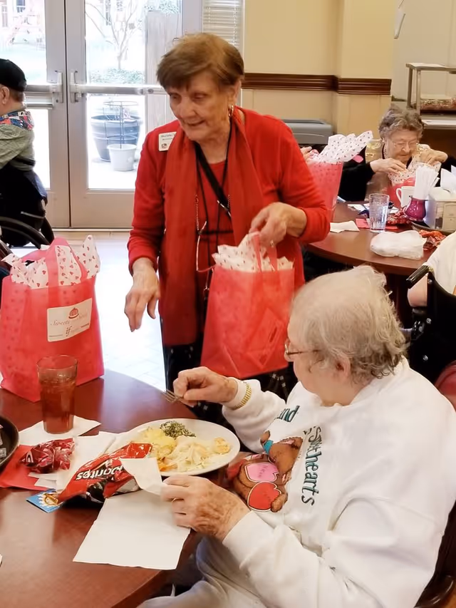 An elderly woman in a white sweatshirt is seated at a table eating a meal with a plate of food and a glass of iced tea. Another elderly woman wearing a red cardigan stands nearby holding a pink gift bag with tissue paper. Other elderly individuals are visible in the background seated at tables in a communal dining area with large windows and doors leading outside.