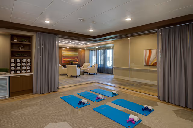 A fitness or exercise room with four blue yoga mats laid out on the floor, each with small dumbbells and towels. The room has a large mirror on one wall, gray curtains on either side, and a seating area with beige armchairs and a colorful abstract painting visible through the mirror reflection.