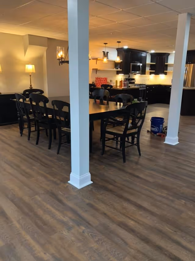 Open dining area with a long dark wooden table and chairs in front of a kitchen with dark cabinets and pendant lighting.