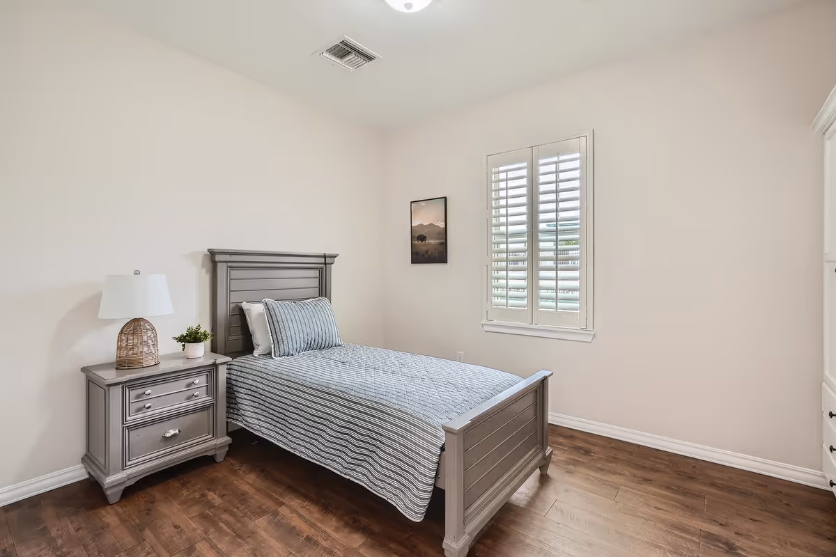 A simple bedroom with a single gray wooden bed with striped bedding, a matching nightstand with a lamp and a small plant, a window with white shutters, a framed picture on the wall, and wooden flooring.