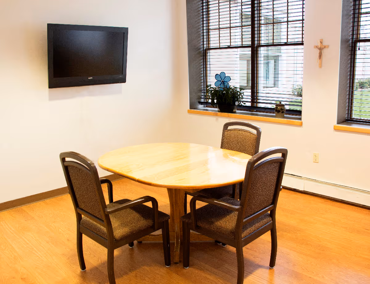 A small room with a wooden table and four chairs arranged around it. A flat-screen TV is mounted on the wall to the left. Two windows with blinds are on the right wall, with a potted plant and decorative items on the windowsill. A wooden crucifix is mounted on the wall between the windows.