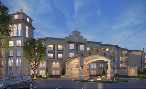 Exterior view of a multi-story senior living facility building at dusk with a covered entrance, arched windows, and a parking area with a car in front.