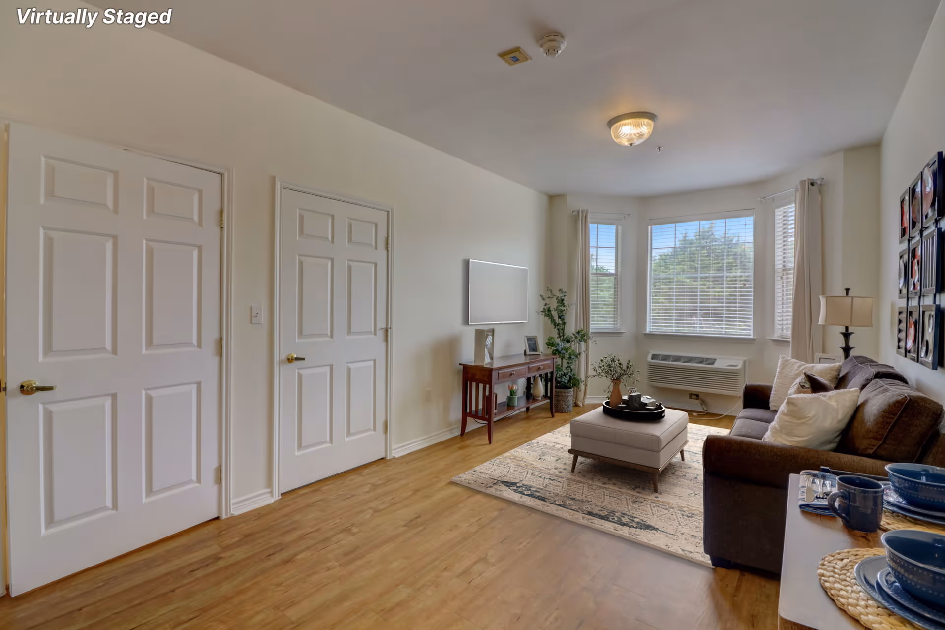 A bright living room with wooden flooring, a brown sofa with white and brown pillows, a beige ottoman on a patterned rug, a wooden console table with decorative items, and a large window with white blinds and curtains letting in natural light. Two white doors are visible on the left wall.