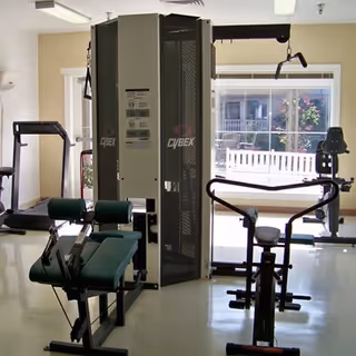 A well-lit fitness room with various exercise equipment including a Cybex multi-station machine, a stationary bike, and a treadmill. Large windows in the background provide natural light and a view of an outdoor area with a white fence and greenery.