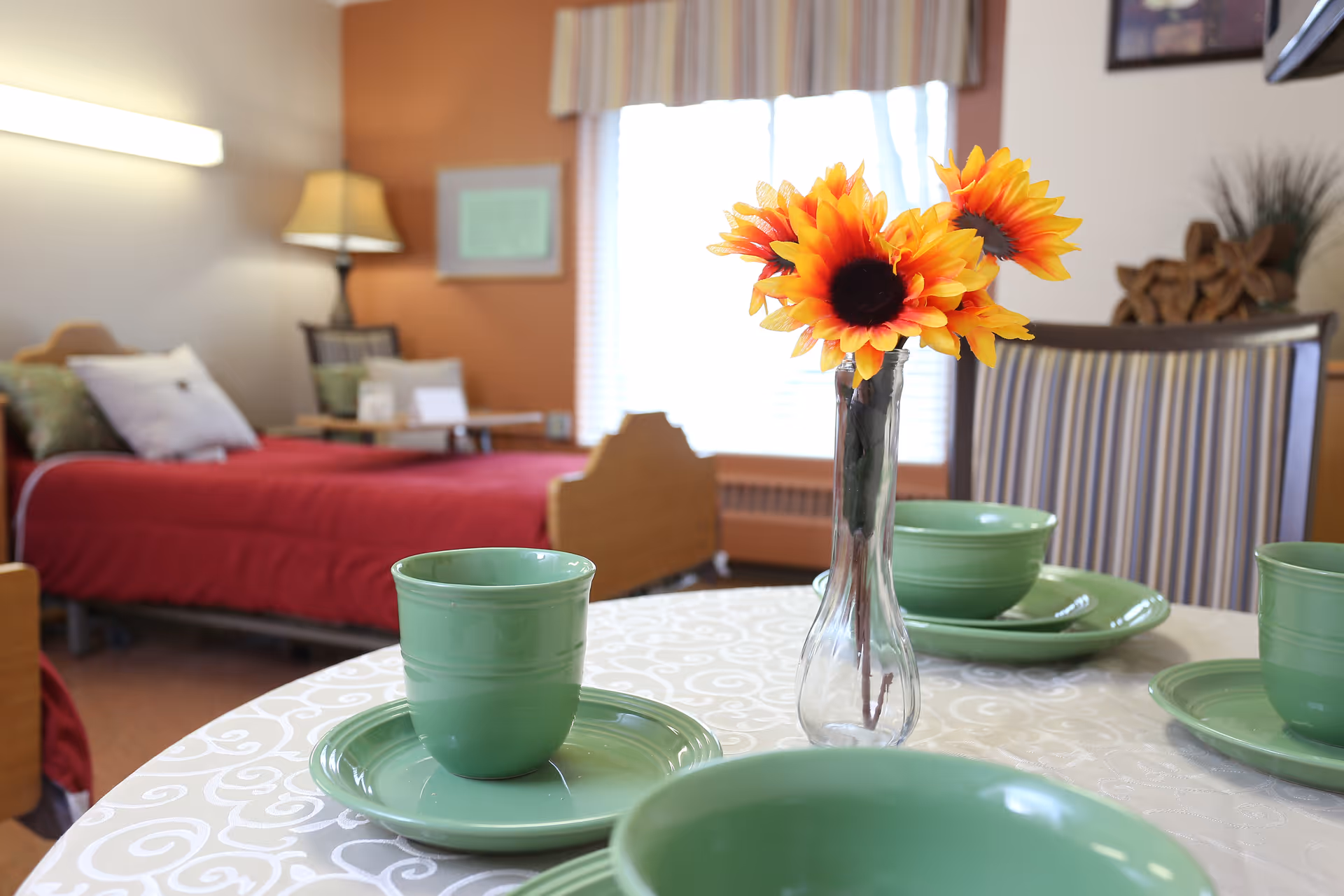 A cozy room with a bed covered in a red blanket in the background and a table set with green dishes and cups in the foreground. A clear vase with bright orange sunflowers is placed on the table. The room has warm lighting and a window with striped curtains.