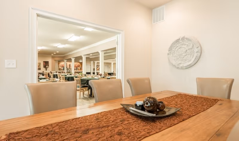 Wood dining table with a decorative runner and centerpiece in the foreground looking into a large dining room with multiple tables and chairs.