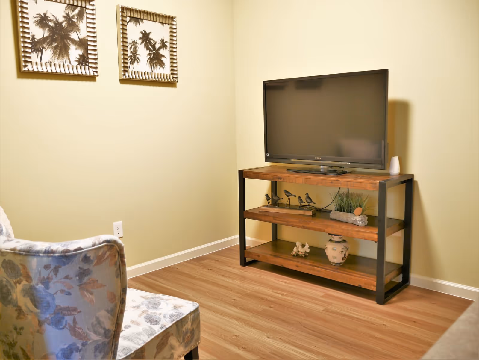 A cozy living room corner with a flat-screen TV on a wooden and metal stand. The stand has decorative items including bird figurines, a small plant, and a vase. A floral upholstered chair is partially visible on the left side. Two framed pictures of palm trees hang on the light yellow wall above the chair.