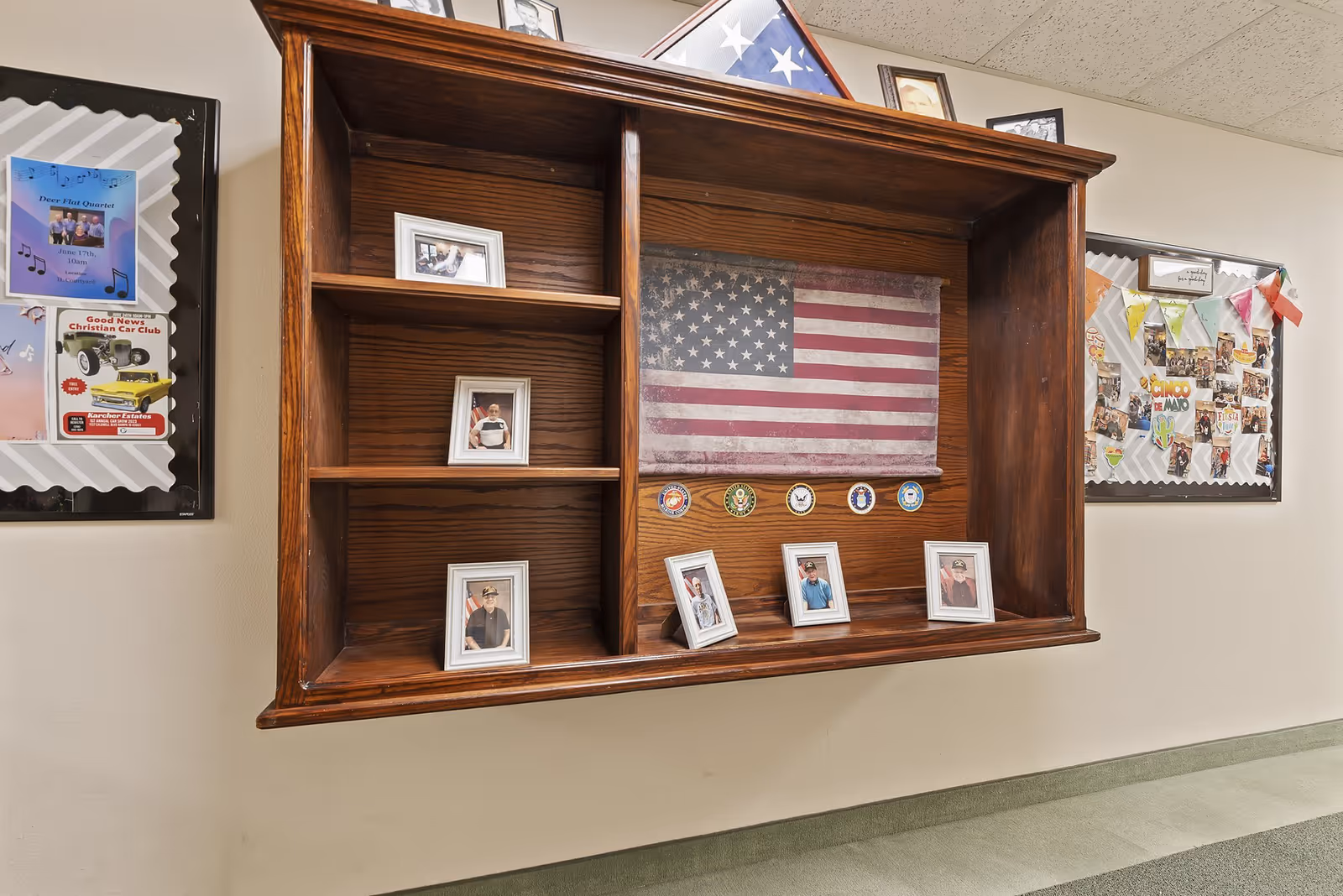 A wooden wall-mounted display case featuring a folded American flag, military service emblems, and framed photographs of veterans. On either side of the display case are bulletin boards with various flyers, photos, and decorations.