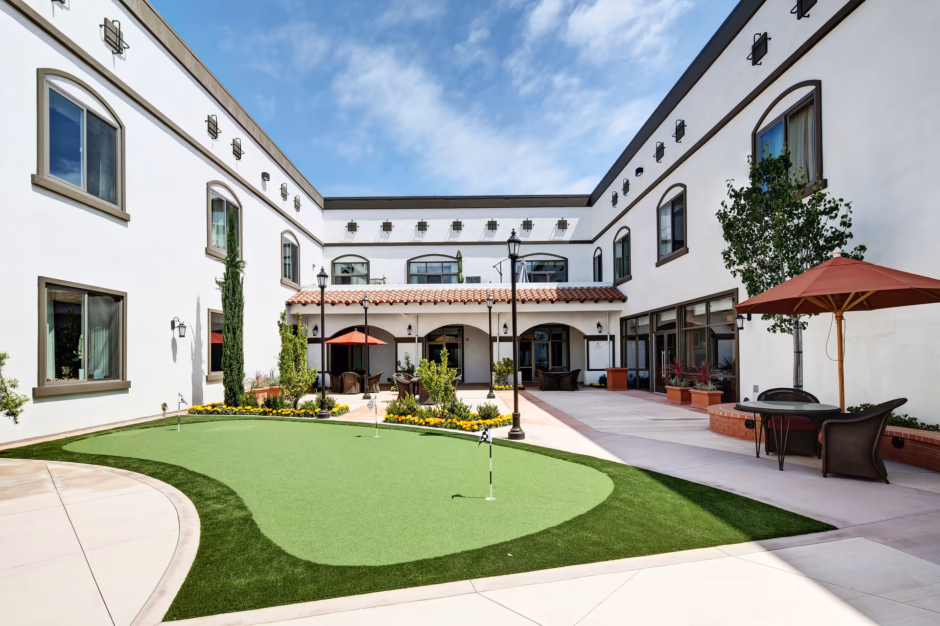 Outdoor courtyard area of Clearwater at South Bay featuring a small putting green with flags, surrounded by a paved walkway. There are patio tables with umbrellas and chairs, decorative plants, and a two-story white building with multiple windows and outdoor lighting fixtures under a partly cloudy sky.