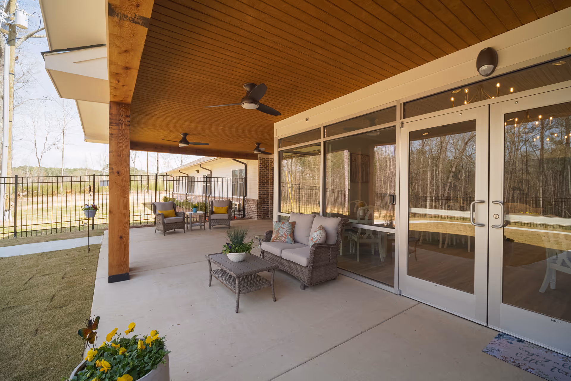 Covered outdoor patio area with wicker seating including a loveseat and chairs, a coffee table with a plant, ceiling fans, and glass doors leading inside. The patio overlooks a fenced yard with trees in the background.