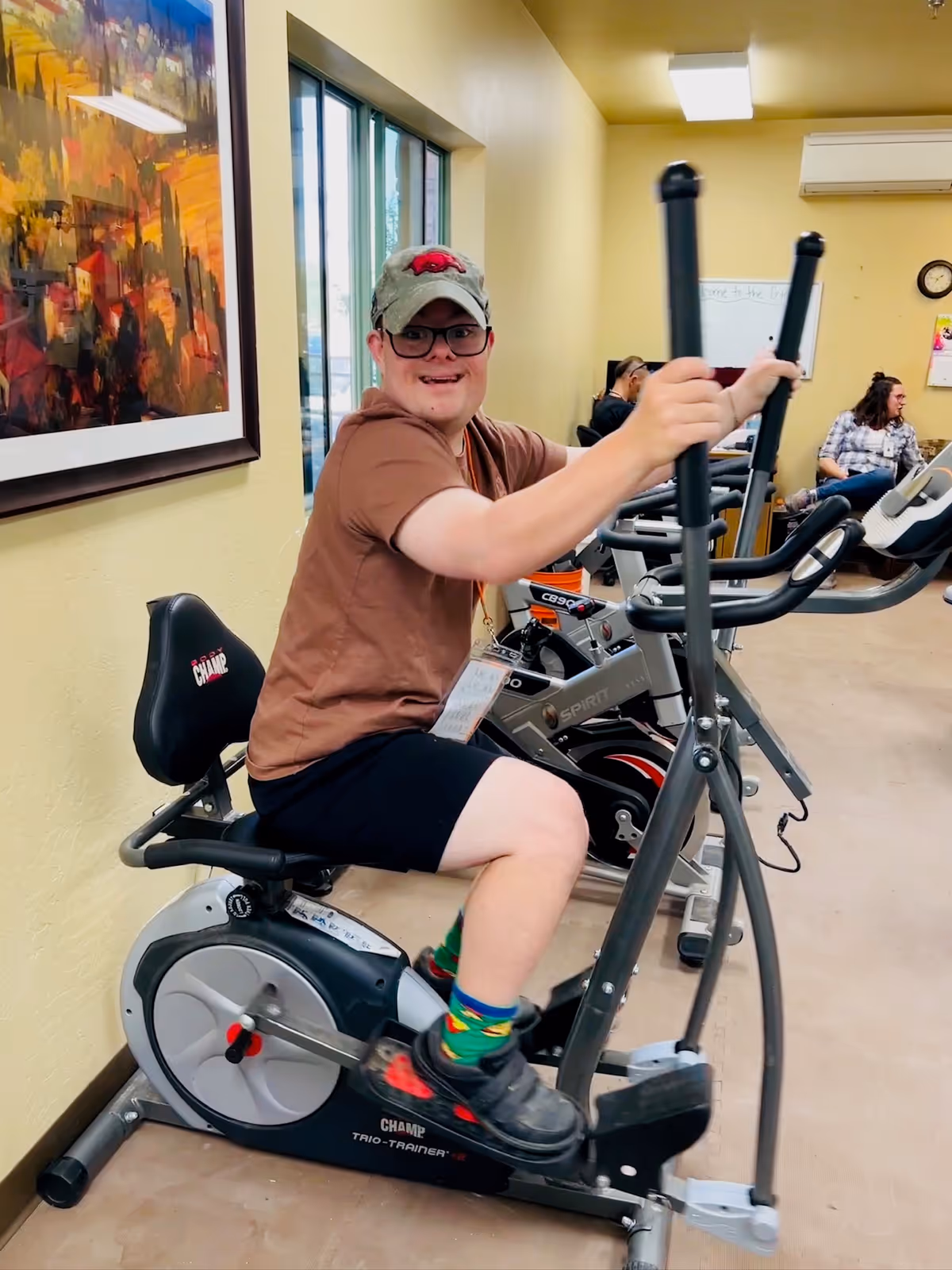 A young man wearing a brown shirt, black shorts, colorful socks, and a cap is smiling while using an elliptical exercise machine in a fitness room. Behind him, there are other exercise bikes and two people sitting and talking in the background. The room has light yellow walls, a large window, a colorful painting on the wall, and a clock.