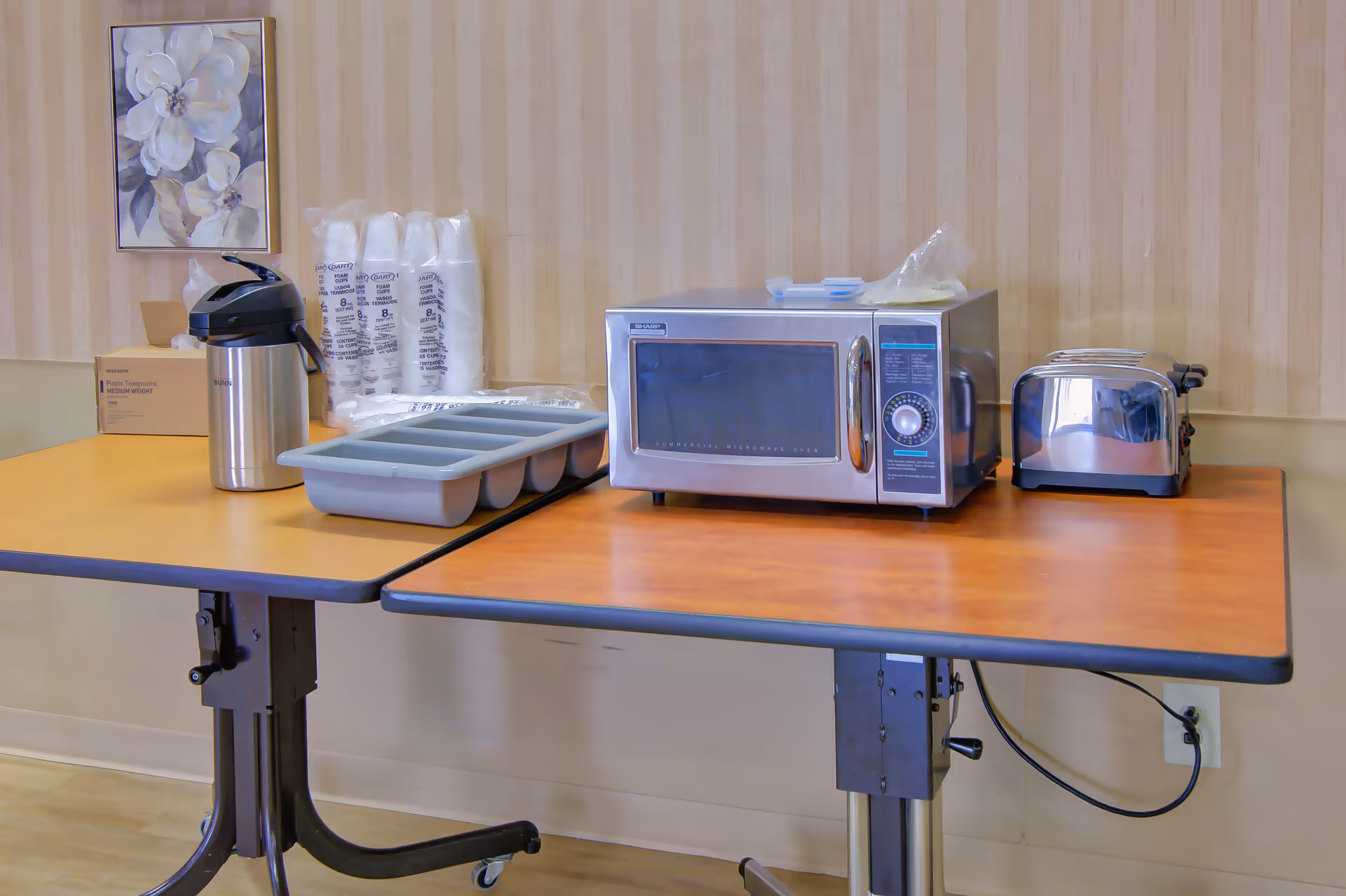 Two tables pushed together with a coffee dispenser, stacks of disposable cups, a gray plastic tray, a microwave oven, and a toaster on top. A floral painting hangs on the striped wallpapered wall behind the tables.