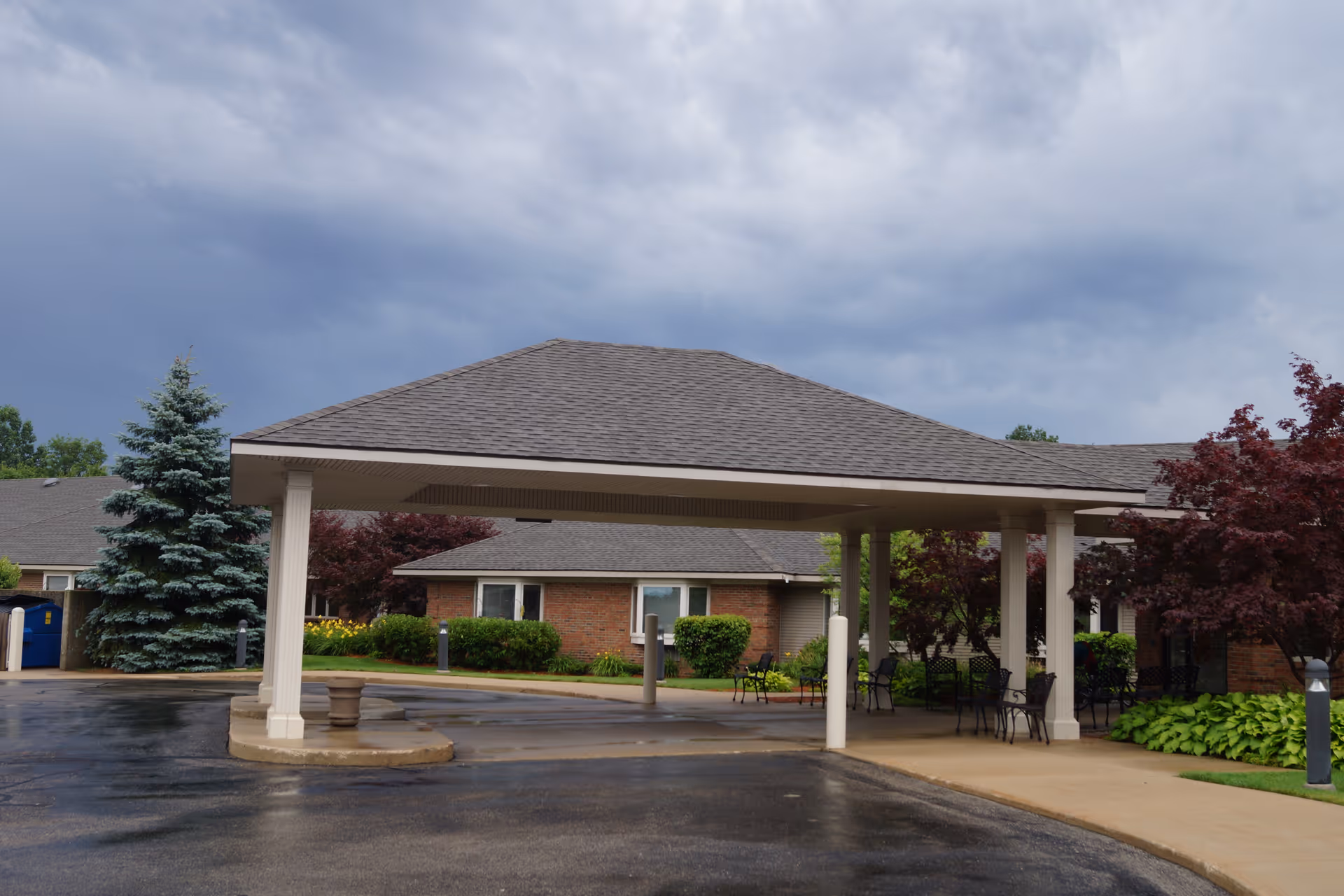 Covered entrance porte-cochere at the front of a brick assisted living building with landscaping and a cloudy sky.