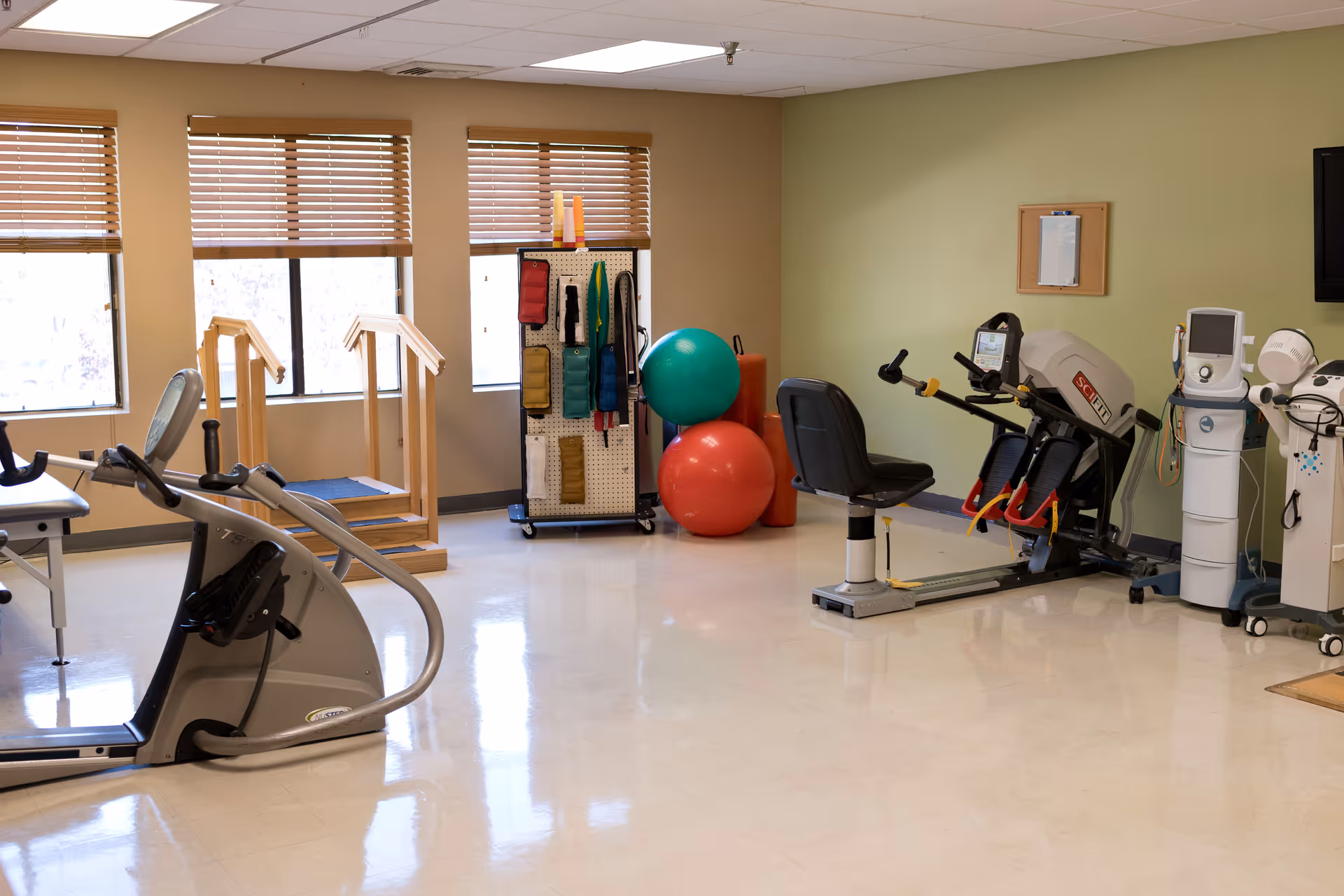 A physical therapy or rehabilitation room with exercise equipment including a stationary bike, parallel bars for walking practice, therapy balls, resistance bands, and various medical devices. The room has large windows with wooden blinds and light-colored walls.