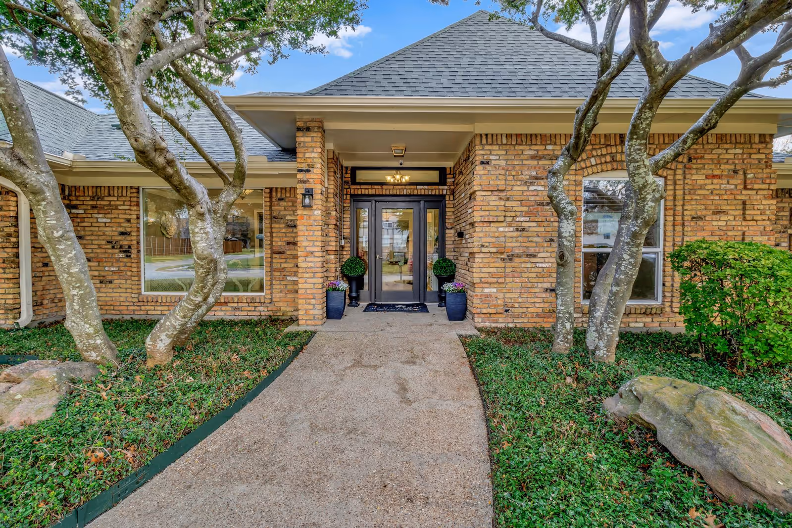 Front entrance of a brick building with a walkway, double glass doors, potted plants, and trees framing the facade.