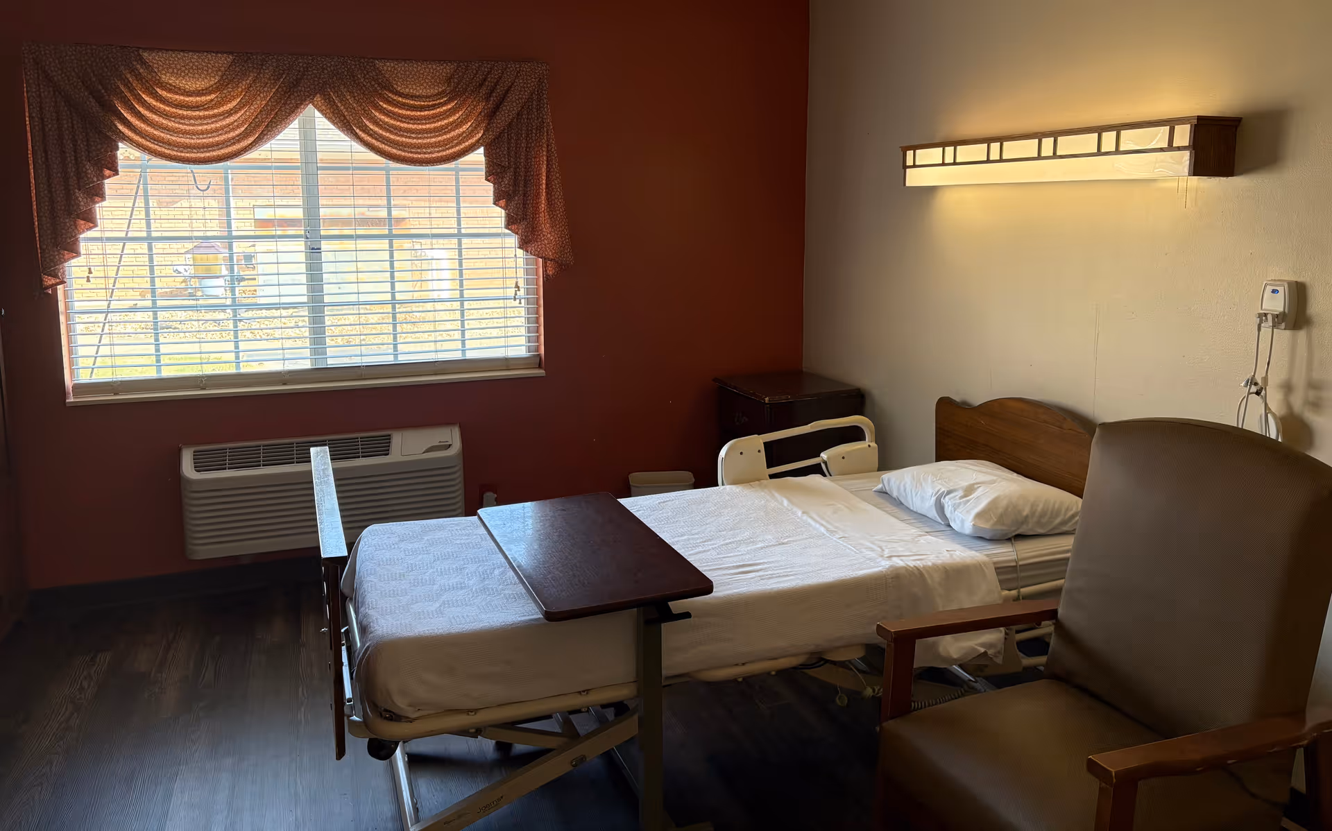 A senior care resident room with a hospital-style bed, overbed table, armchair, bedside cabinet and a window with a valance.