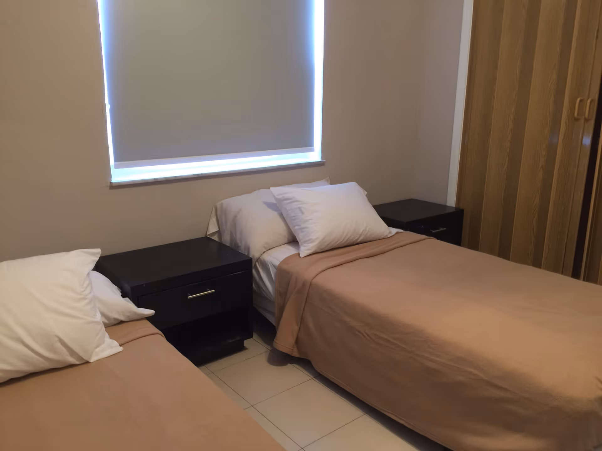 A small bedroom with two single beds, each with a white pillow and beige blanket. Between the beds is a black nightstand with a drawer. A window with a closed blind is above the beds, and a wooden wardrobe is visible on the right side of the room.