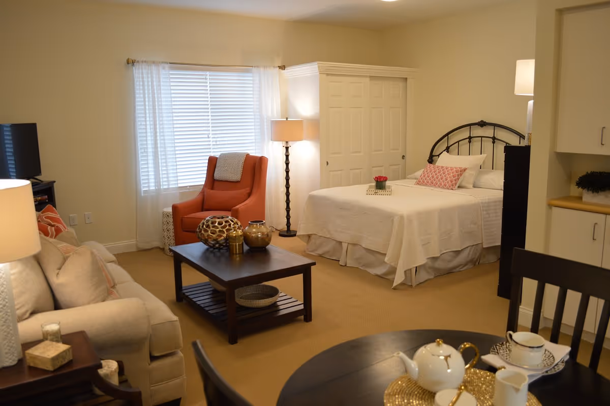 A cozy senior living room and bedroom area featuring a beige sofa with cushions, a wooden coffee table with decorative items, an orange armchair near a window with white blinds and sheer curtains, a bed with white bedding and a pink decorative pillow, a floor lamp, and a small dining table set with a teapot and cups.